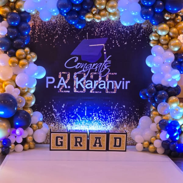 A graduation backdrop with blue, gold, and white balloons, a "Congrats P.A. Karanvir 2025" sign, and "GRAD" boxes.