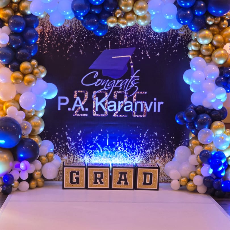 A graduation backdrop with blue, gold, and white balloons, a "Congrats P.A. Karanvir 2025" sign, and "GRAD" boxes.
