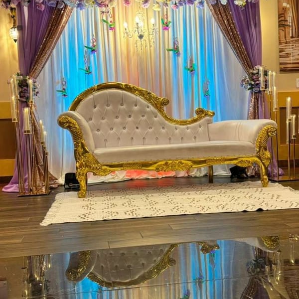 Ornate gold and white chaise lounge on a rug, with purple and gold drapes and blue lighting.