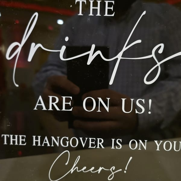 A humorous sign on a dark surface reads, "THE DRINKS ARE ON US! THE HANGOVER IS ON YOU Cheers!"