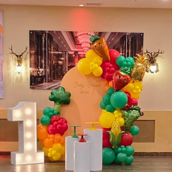 A colorful balloon arch with fruit and vegetable shapes, a large light-up number one, and a sign for "Baby Hrihaan turns 1".