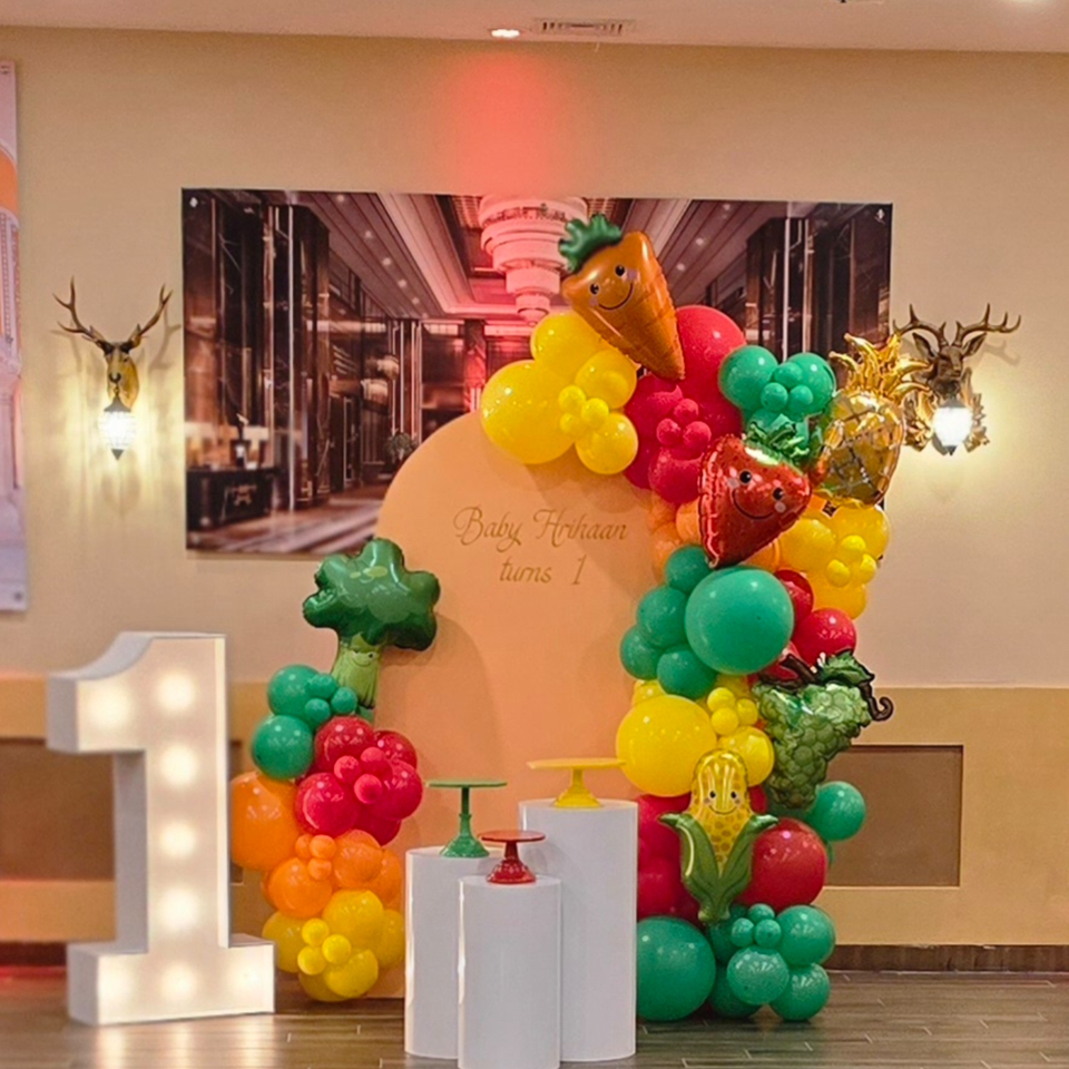 A colorful balloon arch with fruit and vegetable shapes, a large light-up number one, and a sign for "Baby Hrihaan turns 1".
