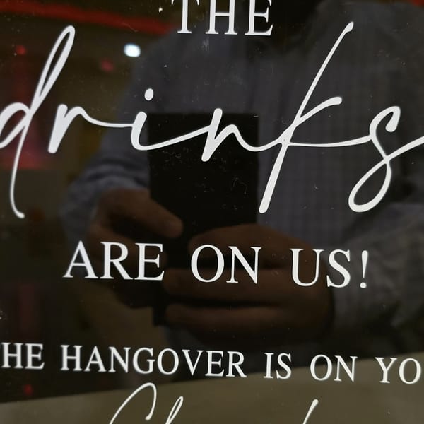 A black sign with white text reading "THE drinks ARE ON US! THE HANGOVER IS ON YOU"