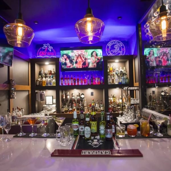 A vibrant bar at Spice Culture with purple lighting, bottles, cocktails, and screens showing dancers.