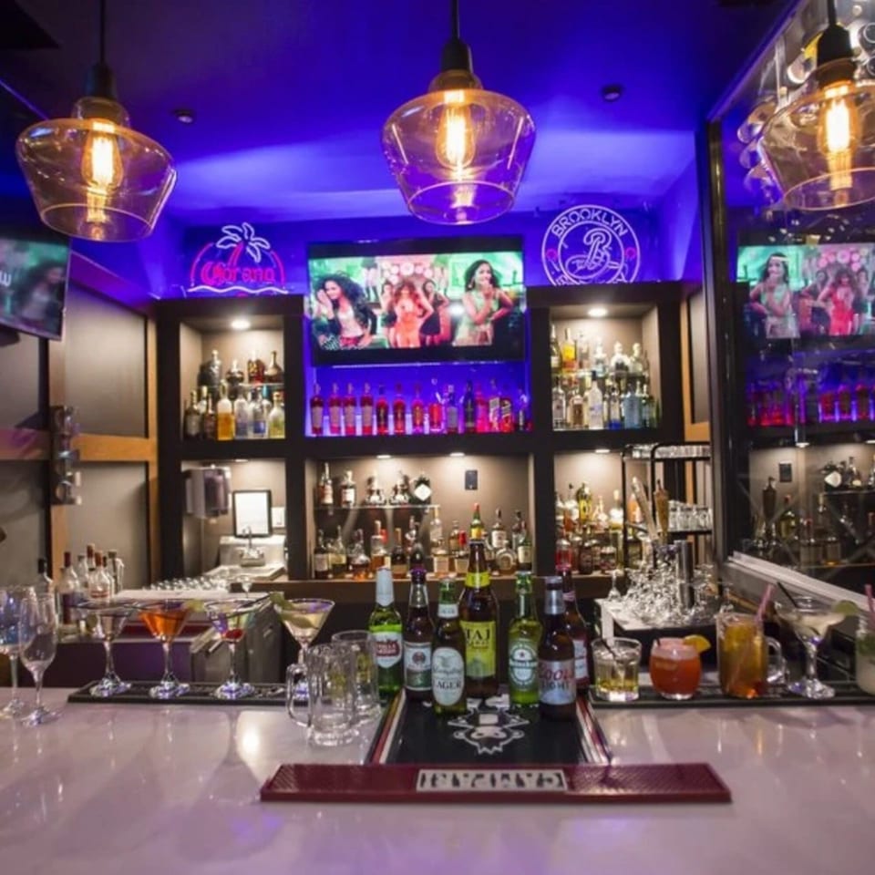 A vibrant bar at Spice Culture with purple lighting, bottles, cocktails, and screens showing dancers.