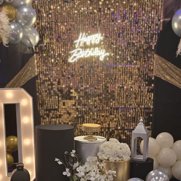 A "Happy Birthday" neon sign glows against a gold sequin wall, with balloons and decorations.