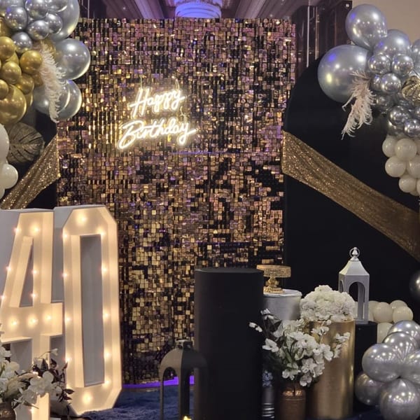 A birthday party setup with a gold sequin backdrop, "Happy Birthday" neon sign, and balloons.