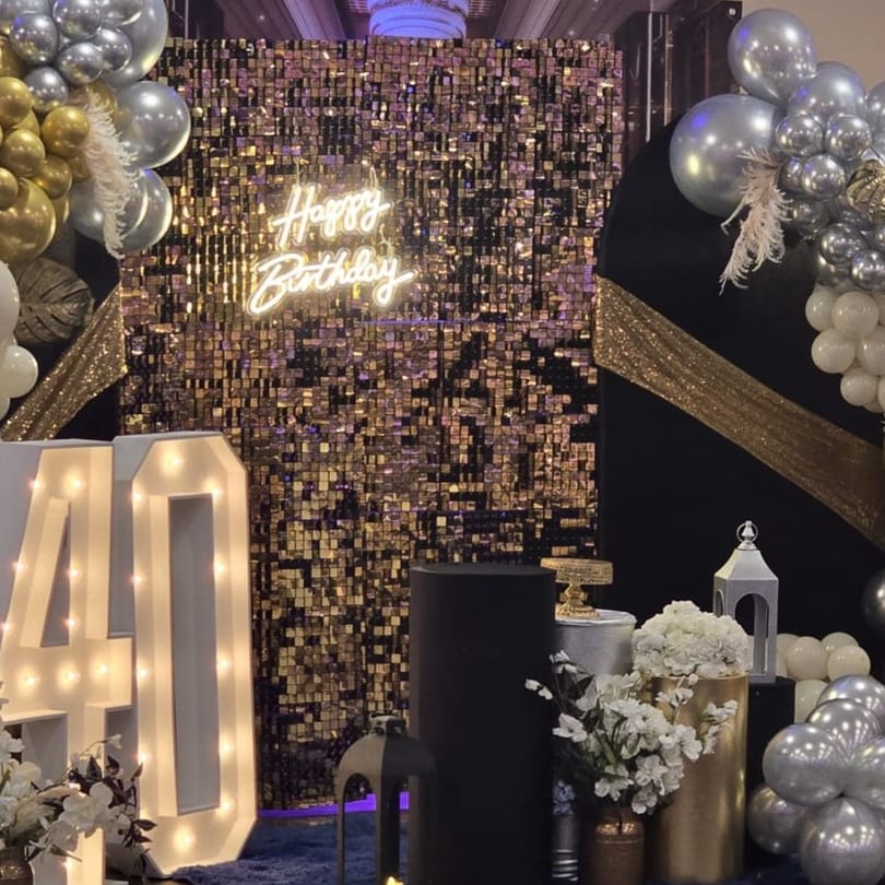 A birthday party setup with a gold sequin backdrop, "Happy Birthday" neon sign, and balloons.