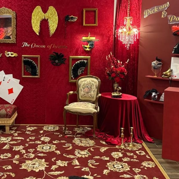 A red-themed photo booth with a vintage chair, masks, golden wings, and playing cards.