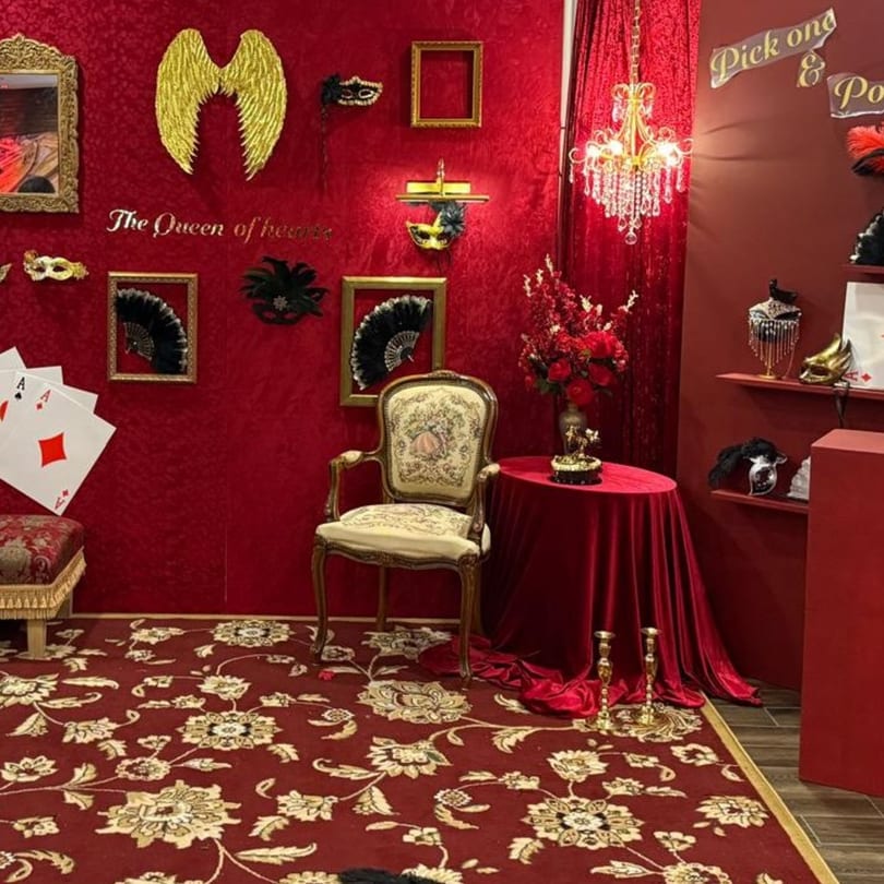 A red-themed photo booth with a vintage chair, masks, golden wings, and playing cards.