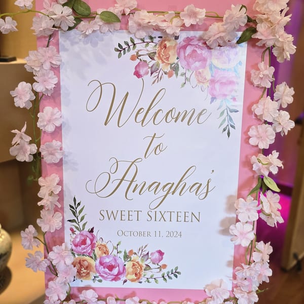 A pink and white welcome sign for "Anagha's Sweet Sixteen" decorated with pink floral garlands.