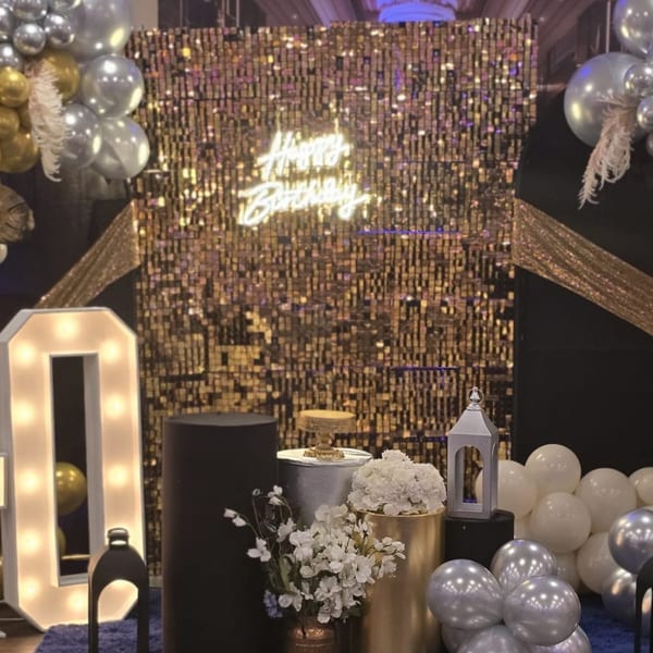 A "Happy Birthday" neon sign glows against a gold sequin backdrop with balloons and floral decor.