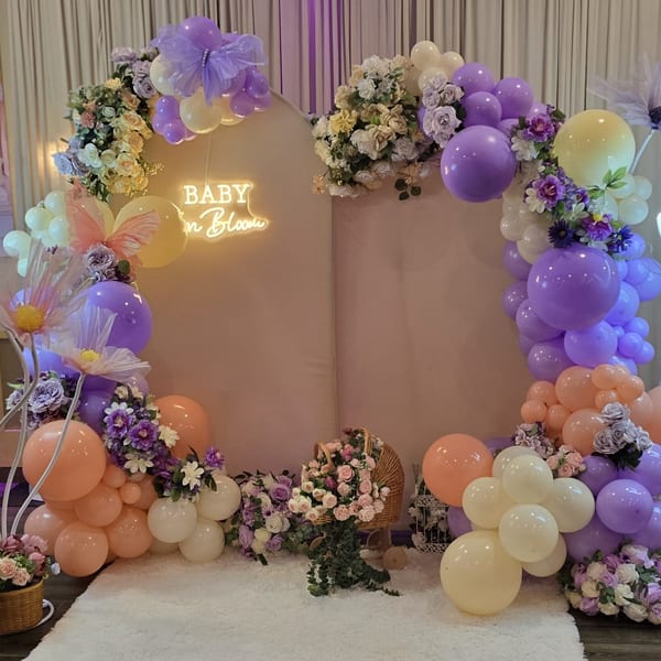 A baby shower backdrop with purple, peach, and cream balloons, flowers, and a "BABY in Bloom" neon sign.
