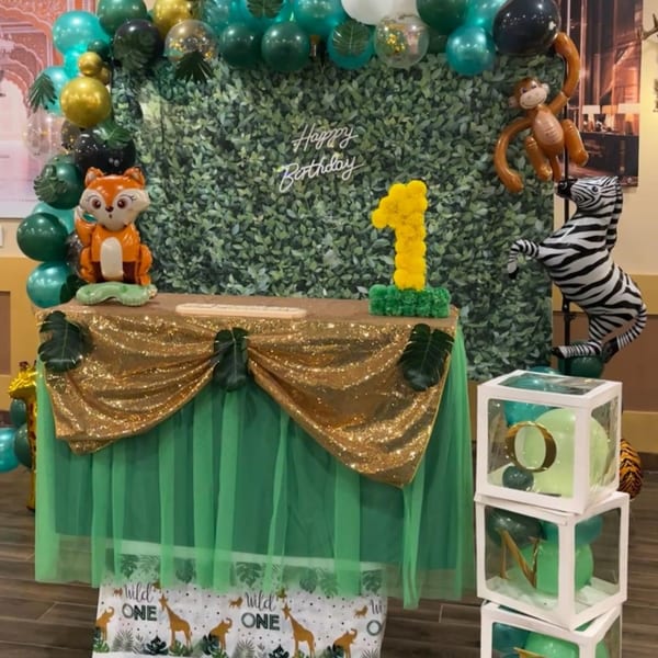 A jungle-themed first birthday party setup with green and gold balloons, animal figures, and a "Happy Birthday" sign.