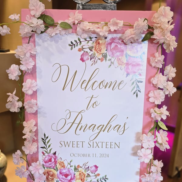 Pink welcome sign for "Anagha's Sweet Sixteen" with floral decorations and a date.