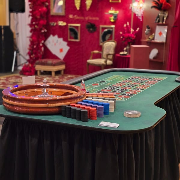 Roulette table with chips and wheel, against a red wall decorated with playing cards and masks.