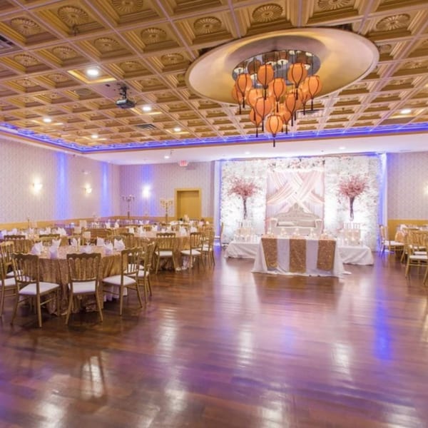 An elegant banquet hall with gold tables, chairs, and a decorated stage, illuminated by blue uplighting.