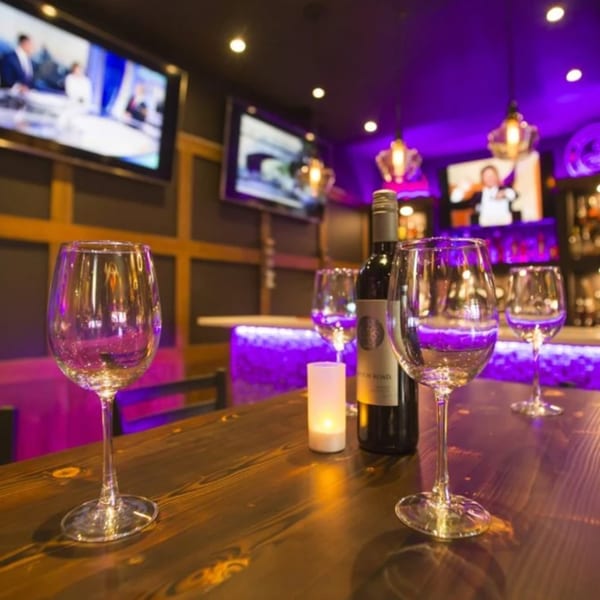 Wine glasses and bottle on a wooden bar with purple lighting and TVs in the background.