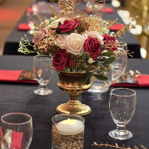 A formal dining table set with a gold vase of red and white roses, glasses, and red napkins.