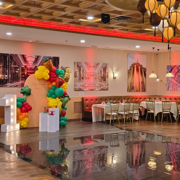 A restaurant dining room with tables, chairs, and a balloon arch for a first birthday celebration.