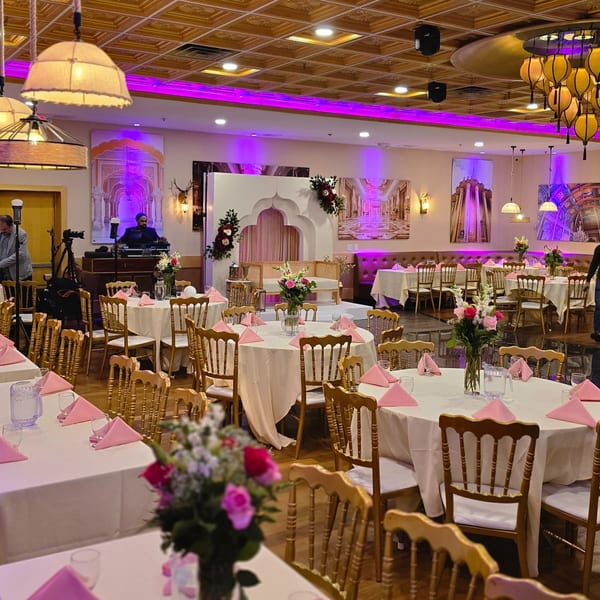 An Indian restaurant event space with round tables, pink napkins, floral centerpieces, and a stage with a sofa.