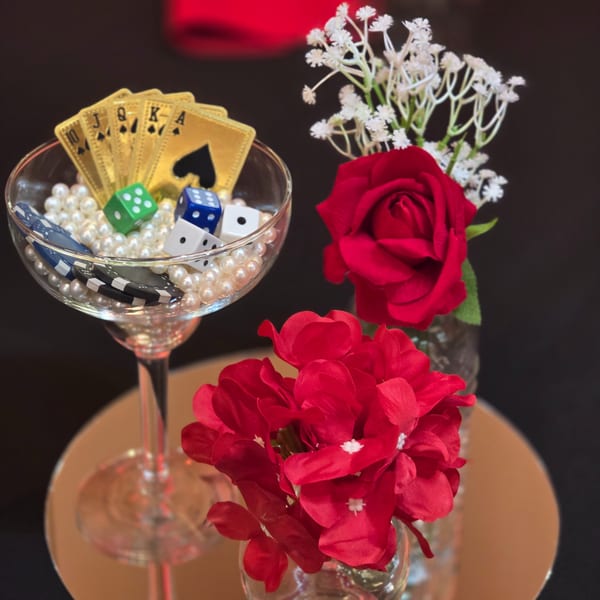 A martini glass filled with pearls, playing cards, dice, and poker chips, next to red roses.