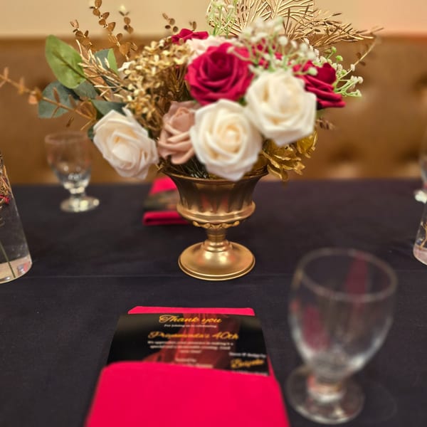 A table setting with a gold vase of red and white roses, glasses, and a "Thank You" card.