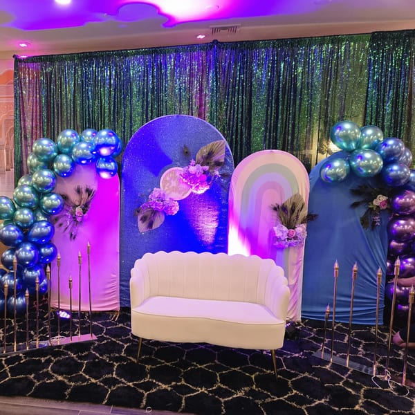 A vibrant event backdrop with blue and purple balloons, peacock feathers, and a white scalloped couch.