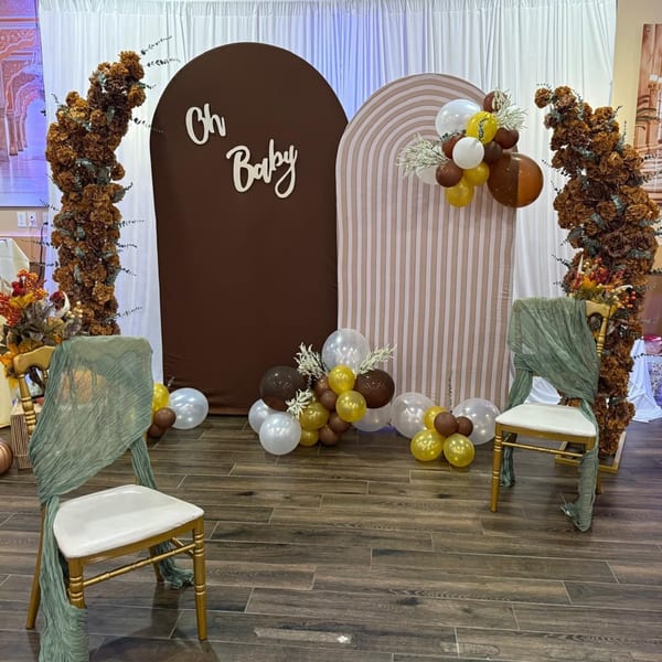 Baby shower decor with "Oh Baby" sign, balloons, floral arches, and two chairs with green sashes.