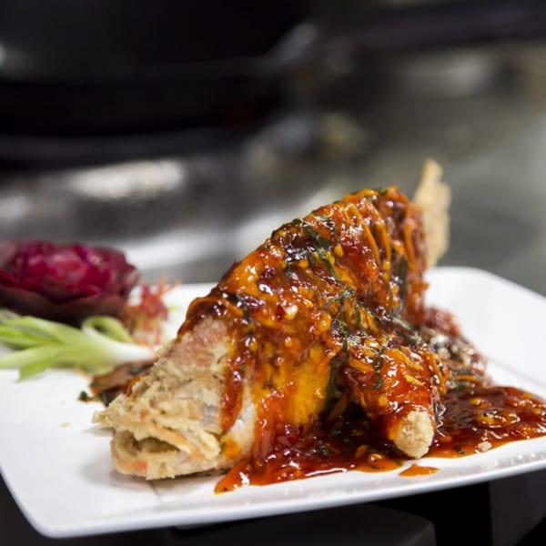 Crispy fried fish covered in a rich, dark orange sauce with green herbs, garnished with red and green vegetables.