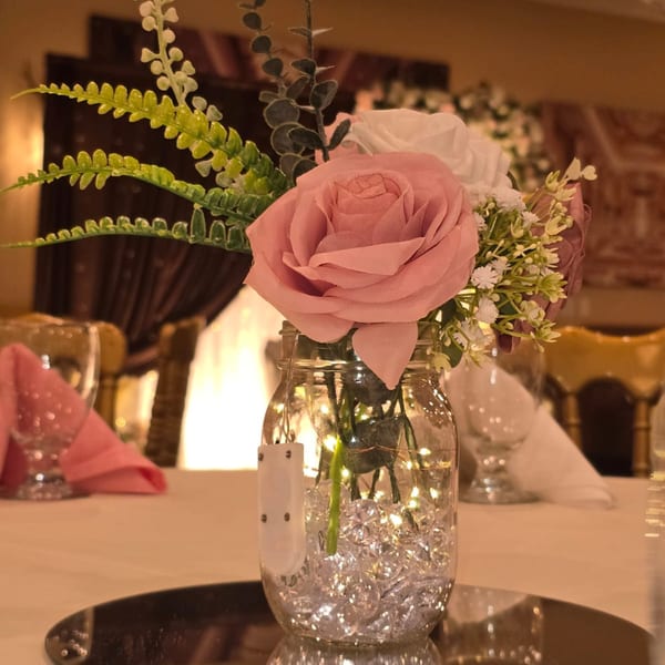 A decorative centerpiece with pink and white flowers in a mason jar with fairy lights on a table.