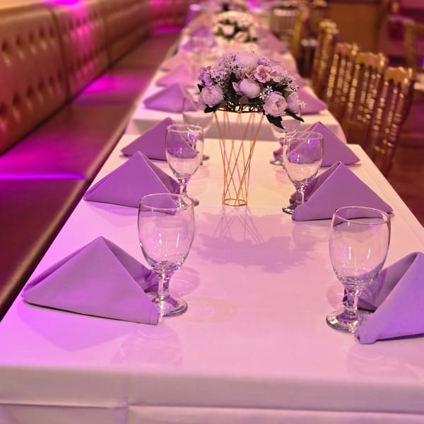Long dining table set with purple napkins, wine glasses, and a floral centerpiece under purple lighting.