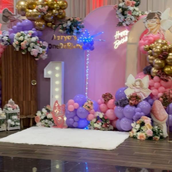 Pink and purple balloon arch with flowers, a light-up '1', and "Happy Birthday" neon sign.