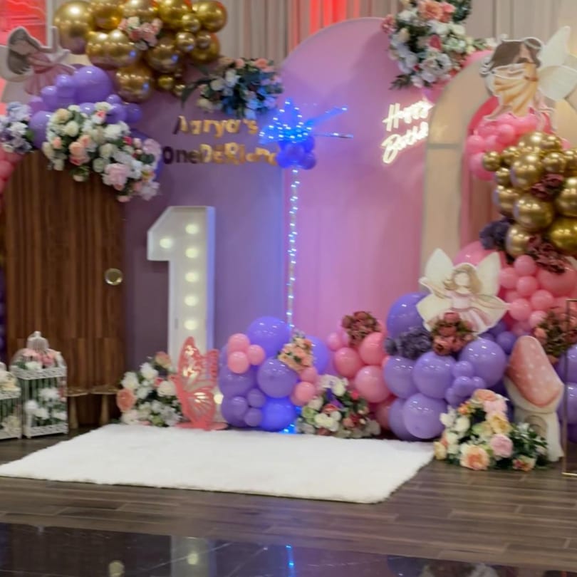 Pink and purple balloon arch with flowers, a light-up '1', and "Happy Birthday" neon sign.