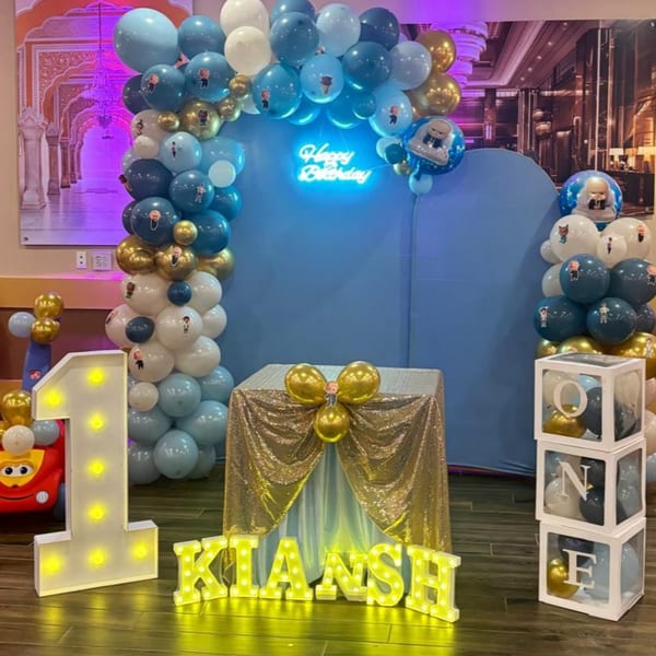 A blue and gold balloon arch with a "Happy Birthday" neon sign, a light-up "1" and "KIANSH" sign.