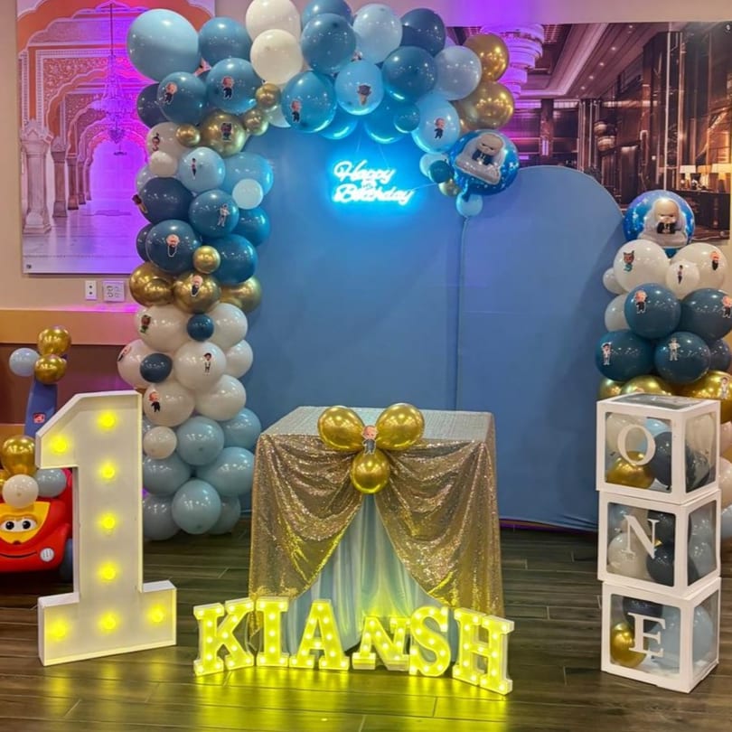 A blue and gold balloon arch with a "Happy Birthday" neon sign, a light-up "1" and "KIANSH" sign.